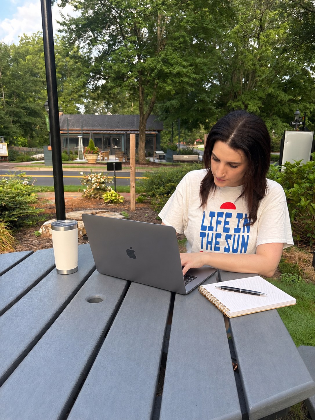 Brittany working on her laptop at an outdoor table, notebook and pen beside her.