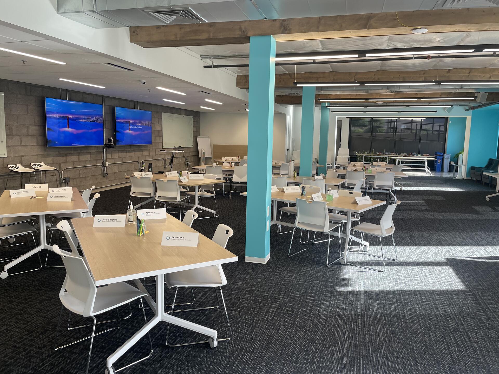 A classroom prepared for instructor-led training with grouped tables, participant name cards, and presentation screens.